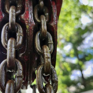 K&ouml;nnte beinhalten: Nahaufnahme eines verwitterten, rot-braunen Metallkettenzugs mit dicken, dunkelbraunen Ketten. Der Flaschenzug ist teilweise sichtbar, wobei die Ketten herunterh&auml;ngen. Der Hintergrund ist verschwommen und zeigt gr&uuml;nes Laub.