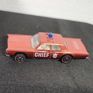 May include: A red toy car with the word "CHIEF" in white lettering on the side. The car has black wheels and a blue light on the roof. The words "FIRE DEPT" are printed on the back of the car.