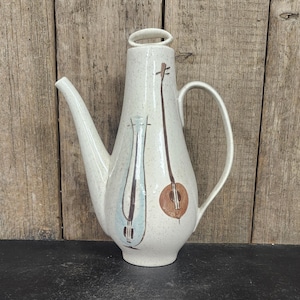 May include: A speckled, off-white ceramic pitcher with a long spout and handle. The pitcher is decorated with two stylized musical instrument designs in brown and blue. The pitcher is likely for serving beverages.