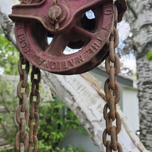K&ouml;nnte beinhalten: Ein antikes rotes Flaschenzugsystem mit Kette, mit der Aufschrift "THE NATIONAL SCREW MFG. CO." und "C-4 1/2". Die Flaschenzug ist im Freien aufgeh&auml;ngt, mit einem verschwommenen Hintergrund aus B&auml;umen und einem Haus.