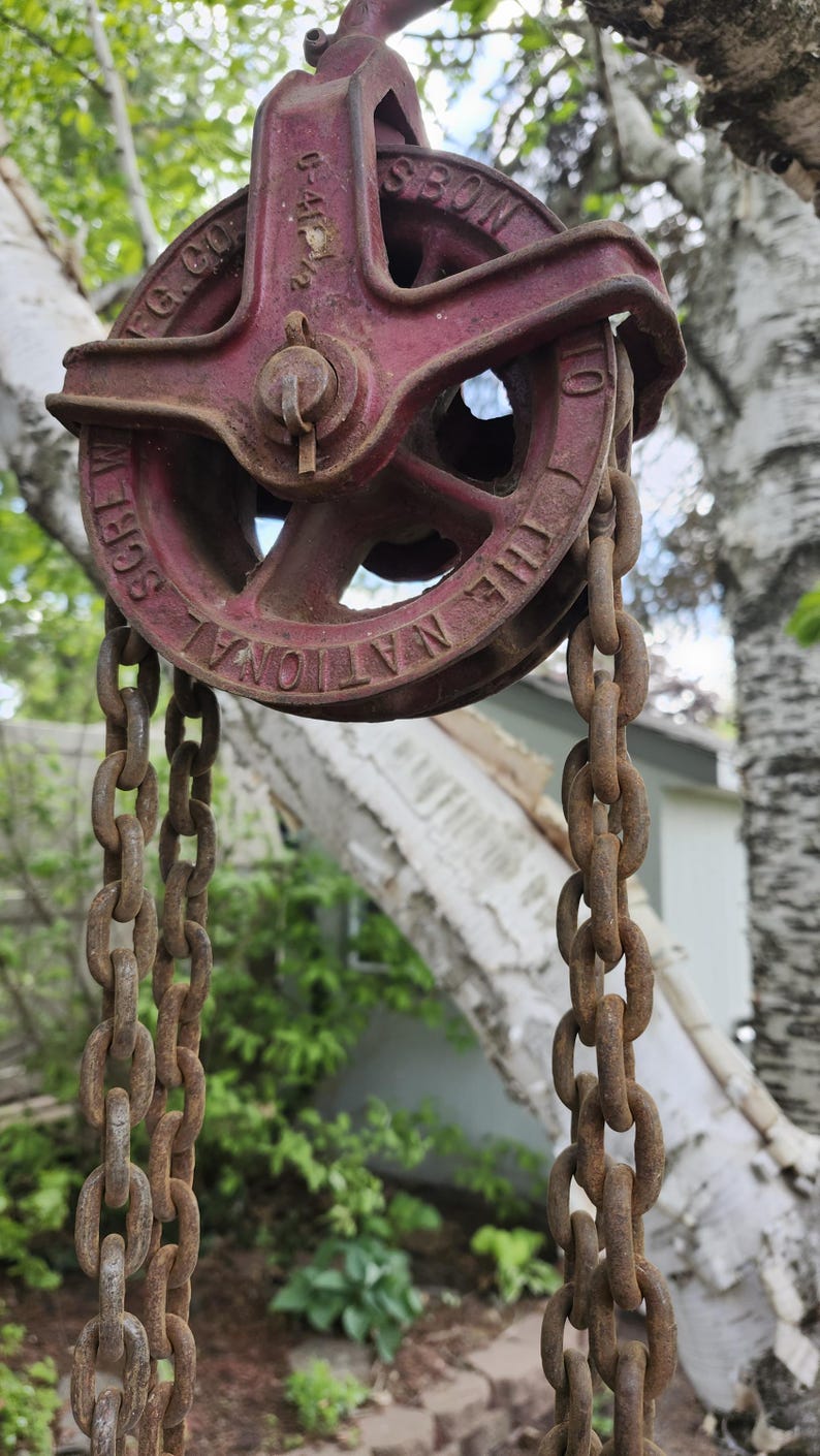K&ouml;nnte beinhalten: Ein antikes rotes Flaschenzugsystem mit Kette, mit der Aufschrift "THE NATIONAL SCREW MFG. CO." und "C-4 1/2". Die Flaschenzug ist im Freien aufgeh&auml;ngt, mit einem verschwommenen Hintergrund aus B&auml;umen und einem Haus.
