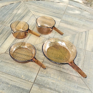 May include: A set of four amber-colored glass cookware pieces consisting of two small saucepans with pouring spouts and two shallow skillets with long handles. The glass is translucent and brown, featuring smooth surfaces and integrated handles.