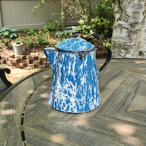 May include: A vintage blue and white enamelware coffee pot with a lid. The pot has a classic speckled pattern and a black handle. It is sitting on a wooden table outdoors.