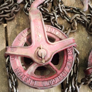 K&ouml;nnte beinhalten: Ein rotes und metallenes Flaschenzugsystem mit einer Kette. Das Flaschenzugrad ist mit "LISBON MFG. CO." und "THE NATIONAL SCREW & MFG. CO." gekennzeichnet. Kette und Flaschenzug befinden sich auf einer Holzoberfl&auml;che.