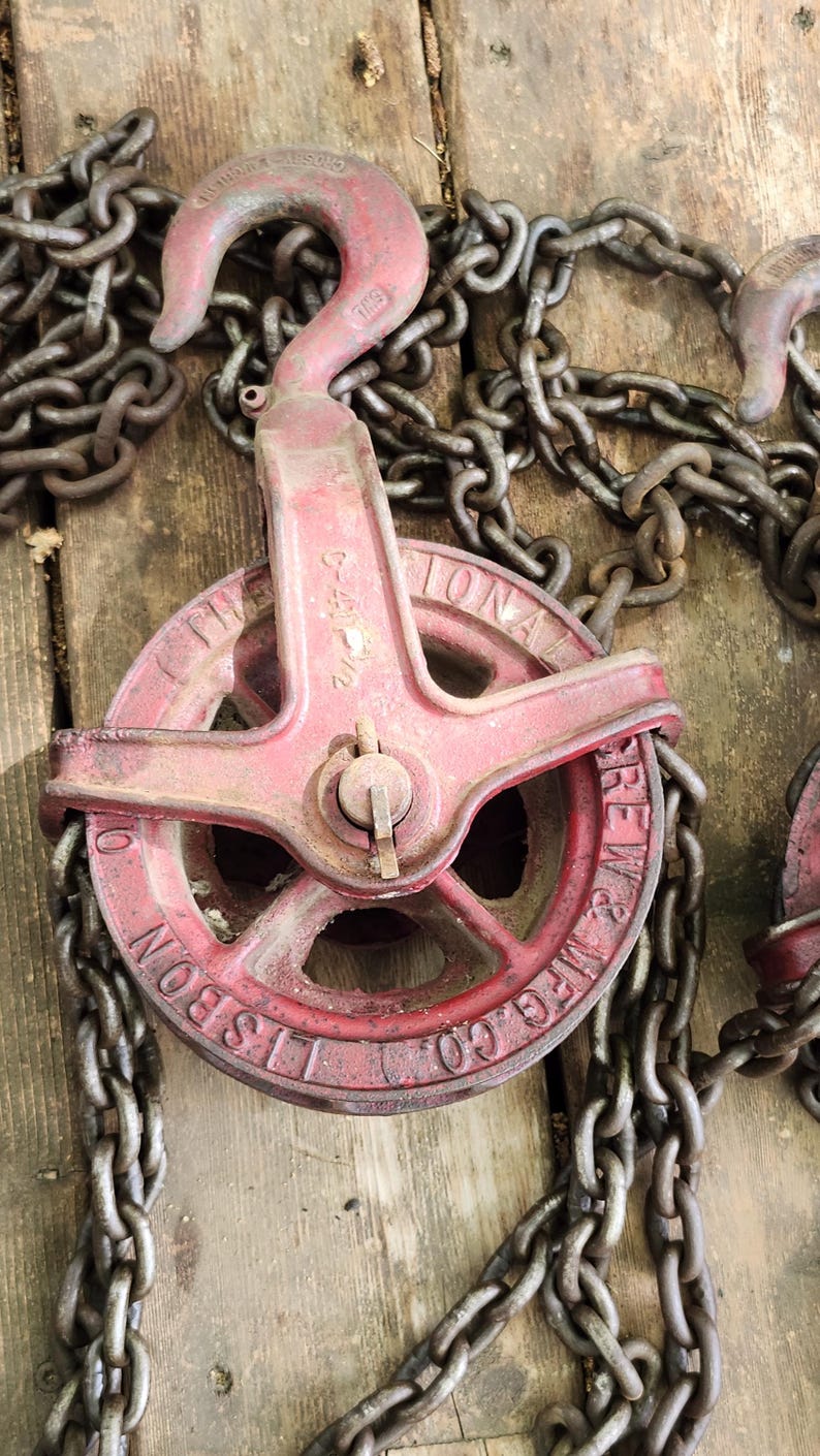 K&ouml;nnte beinhalten: Ein rotes und metallenes Flaschenzugsystem mit einer Kette. Das Flaschenzugrad ist mit "LISBON MFG. CO." und "THE NATIONAL SCREW & MFG. CO." gekennzeichnet. Kette und Flaschenzug befinden sich auf einer Holzoberfl&auml;che.