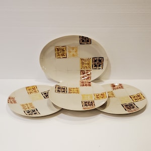 May include: A set of four ceramic plates with a cream-colored background. The plates feature a repeating square pattern in shades of brown, yellow, and black. The set includes one large oval platter and three smaller round plates.