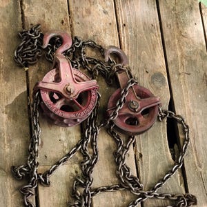 K&ouml;nnte beinhalten: Zwei antike rote Metallrollen mit Ketten, die auf verwitterten Holzbrettern liegen. Die Rollen haben ein kreisf&ouml;rmiges Design mit einem Haken und sind durch eine schwere Kette verbunden. Das Holz hat ein rustikales, gealtertes Aussehen.