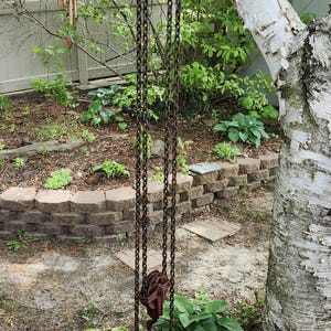 K&ouml;nnte beinhalten: Ein antikes, rostiges rotes Flaschenzugsystem mit einer Kette, das im Freien h&auml;ngt. Die Rolle ist an einem Baum befestigt, wobei die Kette nach unten verl&auml;uft. Der Hintergrund umfasst einen Garten mit Gr&uuml;n und eine Holzterrasse.