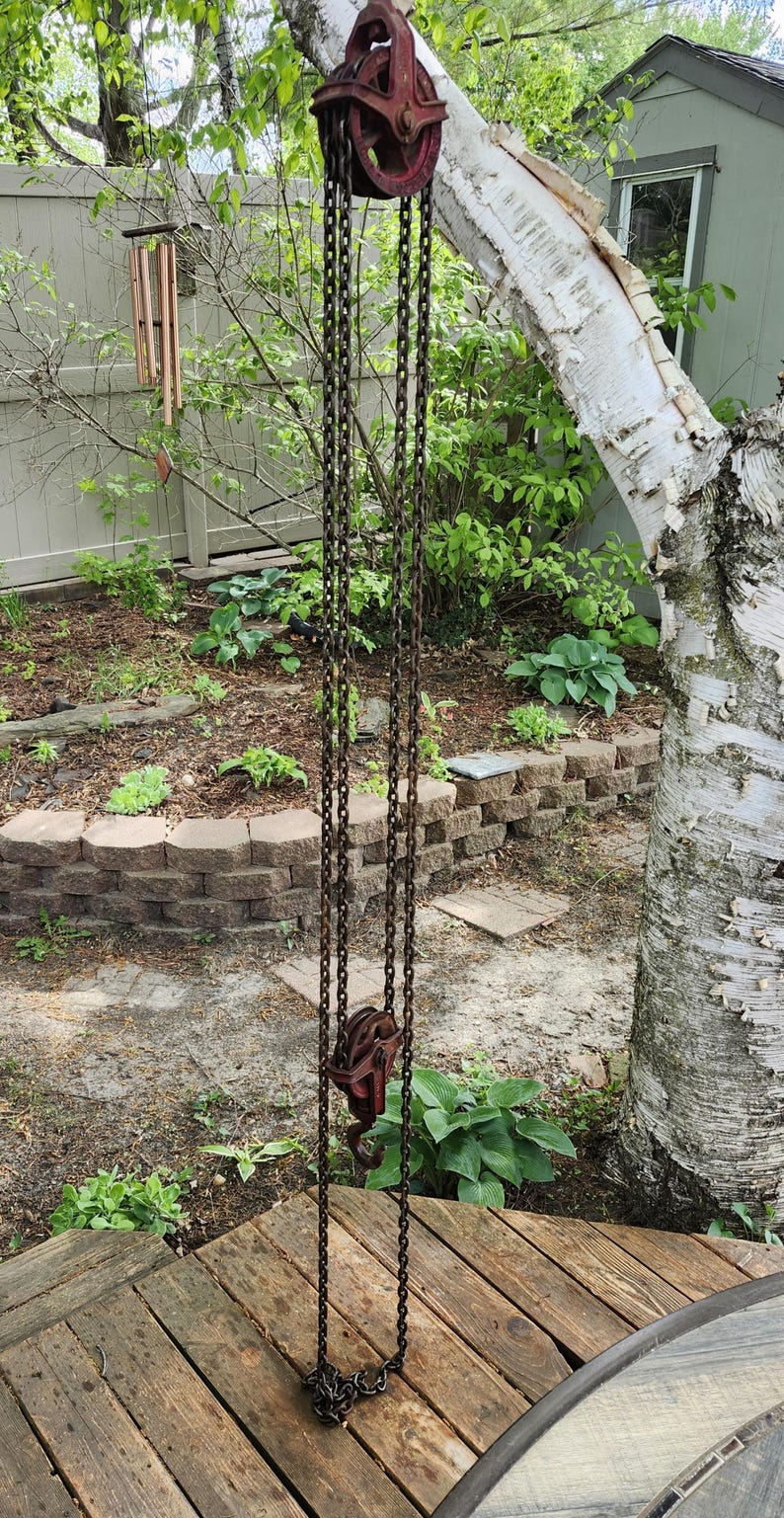 K&ouml;nnte beinhalten: Ein antikes, rostiges rotes Flaschenzugsystem mit einer Kette, das im Freien h&auml;ngt. Die Rolle ist an einem Baum befestigt, wobei die Kette nach unten verl&auml;uft. Der Hintergrund umfasst einen Garten mit Gr&uuml;n und eine Holzterrasse.