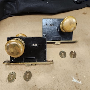 May include: Two antique brass door knobs with black metal plates and keyholes. The knobs are round and have a worn finish. There are four brass keyhole covers on the table.