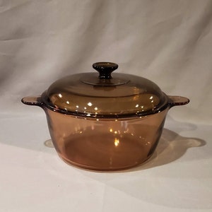 May include: A round, amber-colored glass casserole dish with a matching lid. The dish features two integrated side handles and a central knob on the lid. The glass is translucent, allowing light to pass through, and has a smooth, polished finish.