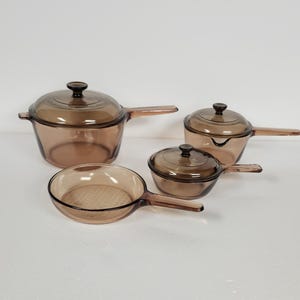 May include: A set of four brown glass cookware pieces, including two pots with lids and a frying pan. The cookware has a classic design with long handles and is suitable for cooking.