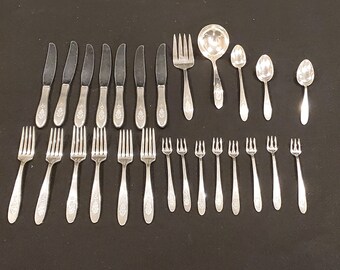 May include: A collection of silver-colored flatware, including knives, forks, spoons, and a ladle, arranged on a dark surface. The utensils have decorative handles with a floral or crest design. The set includes various sizes of forks and spoons.