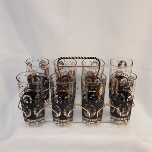 May include: Set of eight clear glass tumblers with a black and gold design. The glasses are arranged in a gold metal rack with a black handle.
