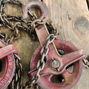 K&ouml;nnte beinhalten: Nahaufnahme von zwei antiken roten Metallrollen mit angebrachten Ketten. Die Rollen haben ein Raddesign und sind durch eine Hochleistungskette verbunden. Der Hintergrund ist eine verwitterte Holzoberfl&auml;che.