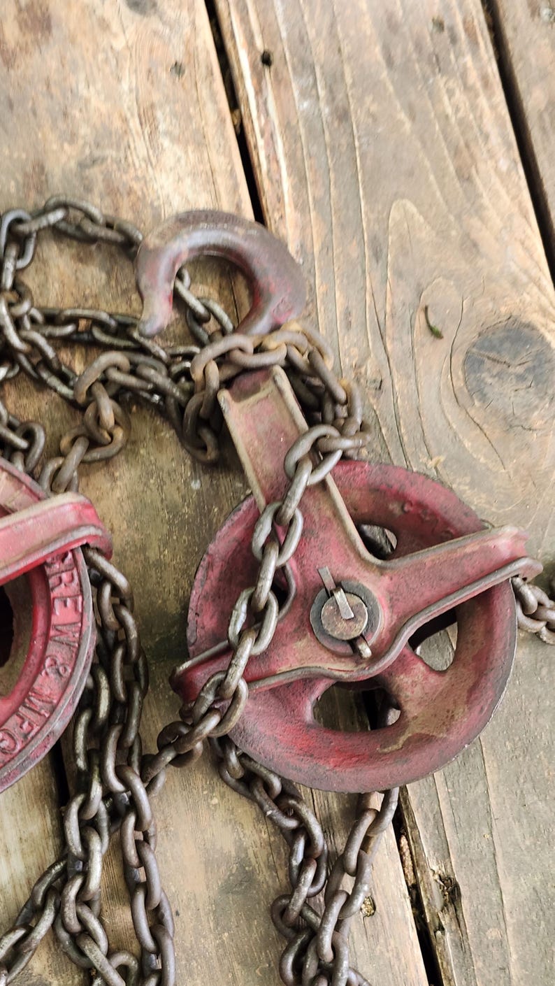 K&ouml;nnte beinhalten: Nahaufnahme von zwei antiken roten Metallrollen mit angebrachten Ketten. Die Rollen haben ein Raddesign und sind durch eine Hochleistungskette verbunden. Der Hintergrund ist eine verwitterte Holzoberfl&auml;che.