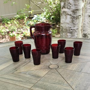 May include: A vintage ruby red glass pitcher and matching glasses set on a gray tiled table. The pitcher has a handle and a flared spout. Eight glasses are arranged around the pitcher. The set is likely for serving beverages.