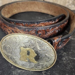 May include: A brown leather belt featuring an intricate floral-patterned embossed design and a dark edge finish. The belt is secured with a large, oval-shaped silver-toned metal buckle that displays detailed scrollwork and a prominent gold-toned letter R.
