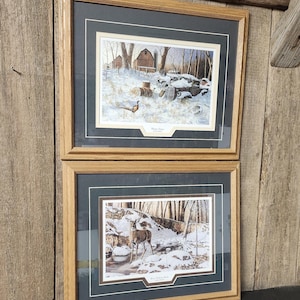 May include: Two framed landscape prints. The top print depicts a snowy scene with a barn and old vehicles. The bottom print shows a deer near a stream in a snowy forest. Both are in wooden frames.