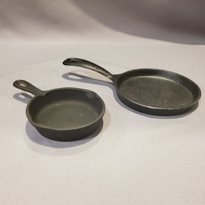 May include: Two black cast iron skillets, one small and one large, on a gray surface.
