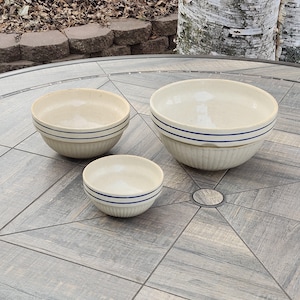 May include: Three ceramic bowls with blue and white stripes. The bowls are nested inside each other, with the largest bowl on the bottom and the smallest bowl on top. The bowls are on a wooden table.