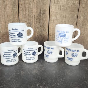 May include: A collection of six white ceramic mugs with various blue text and logos. The mugs feature text such as "Jobber Amoco Products" and "Bergstrom Oil". The mugs are arranged on a dark surface against a wooden background.