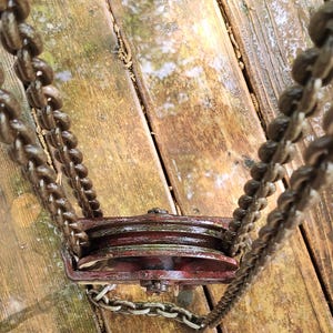 K&ouml;nnte beinhalten: Ein verrostetes, rotes Flaschenzugsystem mit zwei R&auml;dern, verbunden durch eine Kette. Kette und Flaschenzug sind in einem dunklen, verwitterten Braun gehalten, was auf Alter und Nutzung hindeutet. Der Flaschenzug ist auf einer Holzoberfl&auml;che montiert.