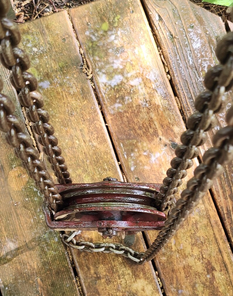 K&ouml;nnte beinhalten: Ein verrostetes, rotes Flaschenzugsystem mit zwei R&auml;dern, verbunden durch eine Kette. Kette und Flaschenzug sind in einem dunklen, verwitterten Braun gehalten, was auf Alter und Nutzung hindeutet. Der Flaschenzug ist auf einer Holzoberfl&auml;che montiert.