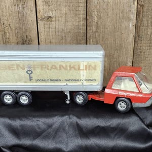 May include: A red toy truck with a gray trailer. The trailer has the words "Ben Franklin" and "Locally Owned - Nationally Known" printed on it.