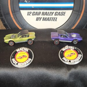 May include: Two vintage Hot Wheels toy cars, one lime green and one purple, with black tops and red-line tires. The cars are displayed with two Hot Wheels logo badges and a Hot Wheels 12 Car Rally Case by Mattel.