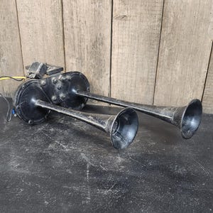 May include: A pair of vintage black metal horns with a trumpet-like shape. The horns are mounted on a black base with wiring and a small electrical component. The horns are likely for a vehicle.