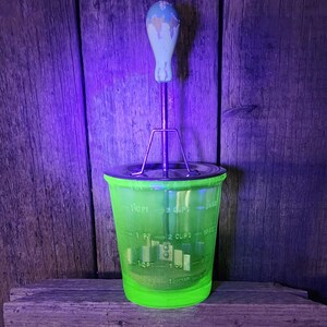 May include: A vintage green glass food mill with a metal blade and a light blue and white handle. The mill has measurement markings on the side and sits on a wooden surface against a wooden background.