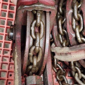 K&ouml;nnte beinhalten: Nahaufnahme eines roten und braunen Kettenzugs im Vintage-Stil. Der Hebezug verf&uuml;gt &uuml;ber eine robuste Kette und einen stabilen Metallrahmen. Die Kettenglieder sind dick und weisen Gebrauchsspuren auf. Das Design deutet auf ein Werkzeug zum Heben schwerer Gegenst&auml;nde hin.