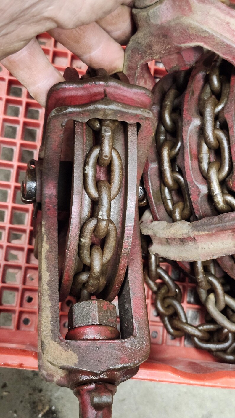 K&ouml;nnte beinhalten: Nahaufnahme eines roten und braunen Kettenzugs im Vintage-Stil. Der Hebezug verf&uuml;gt &uuml;ber eine robuste Kette und einen stabilen Metallrahmen. Die Kettenglieder sind dick und weisen Gebrauchsspuren auf. Das Design deutet auf ein Werkzeug zum Heben schwerer Gegenst&auml;nde hin.