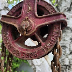 K&ouml;nnte beinhalten: Vintage rote Metallrolle mit Kette. Auf dem Rollenrad sind die Worte "NATIONAL SCREW & MFG CO" und "SBON" eingepr&auml;gt. Kette und Rolle weisen Rostspuren auf.