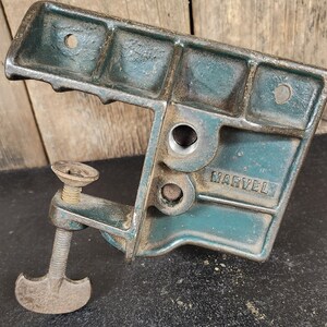 May include: A vintage teal-colored metal clamp with the word "MARVEL" embossed on the side. The clamp features a screw mechanism and a T-shaped handle. The tool is designed for woodworking or metalworking.
