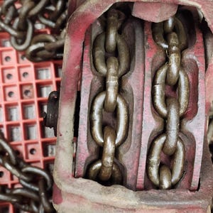 K&ouml;nnte beinhalten: Nahaufnahme eines roten und braunen Kettenzugs im Vintage-Stil. Der Hebezug hat zwei parallele Ketten, die durch ihn verlaufen. Das Metall ist abgenutzt und weist Gebrauchsspuren auf. Der Hintergrund ist ein rotes Kunststoffgitter.