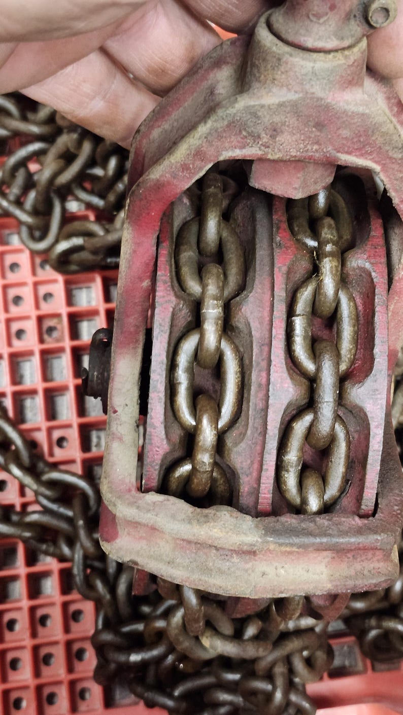 K&ouml;nnte beinhalten: Nahaufnahme eines roten und braunen Kettenzugs im Vintage-Stil. Der Hebezug hat zwei parallele Ketten, die durch ihn verlaufen. Das Metall ist abgenutzt und weist Gebrauchsspuren auf. Der Hintergrund ist ein rotes Kunststoffgitter.