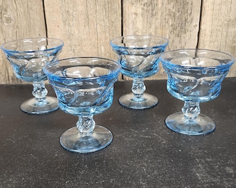 Könnte beinhalten: Vier hellblaue Vintage-Dessertgläser. Jedes Glas hat eine runde Schale mit einem dekorativen Muster und steht auf einem Sockel mit einem gedrehten Stiel. Die Gläser sind auf einer dunklen Oberfläche angeordnet.