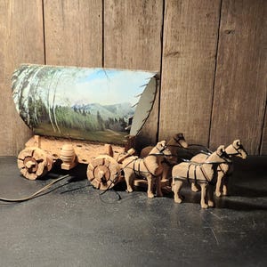 May include: A wooden model of a covered wagon pulled by a team of three carved horses. The wagon features a curved top with a printed landscape scene of mountains and trees. The wheels and horses are crafted from light-colored, textured wood.