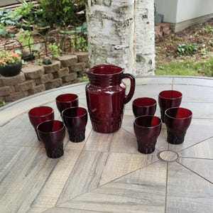 May include: A vintage ruby red glass drinkware set. The set includes a pitcher and eight matching glasses. The pitcher has a handle and a textured pattern. The glasses have a similar design. The set is arranged on a round, patterned table.