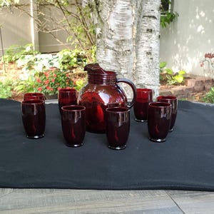 May include: A set of eight ruby red glass tumblers and a matching pitcher. The glasses and pitcher are arranged on a black tablecloth with a wooden table underneath.