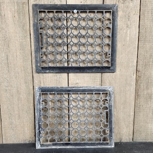 May include: Two rectangular, dark gray metal vent covers with a geometric pattern of interlocking circles. The covers have a rectangular frame and are set against a wooden background.