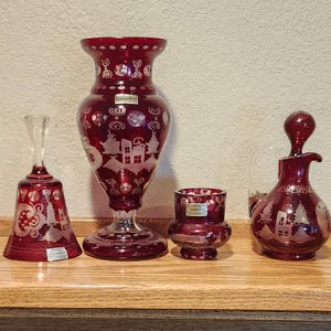 May include: A collection of ruby red glass decorative items, including a bell, a vase, a small cup, and a decanter. Each piece features etched designs of houses and trees. The items are displayed on a wooden surface.