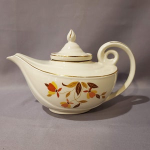 May include: A white teapot with a gold trim and a floral design in shades of orange, yellow, and brown. The teapot has a unique shape with a curved spout and a handle that loops around the top.