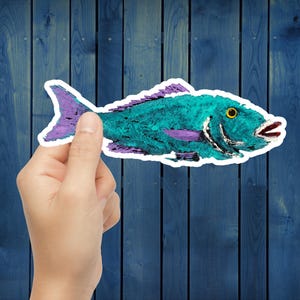 May include: A teal and purple fish sticker with a white border. The fish has a yellow eye and red mouth. The sticker is held against a blue wooden background.