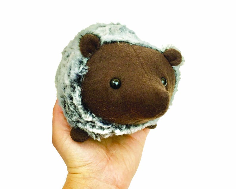 May include: A brown and gray plush hedgehog toy with black eyes. The hedgehog is held in a hand.