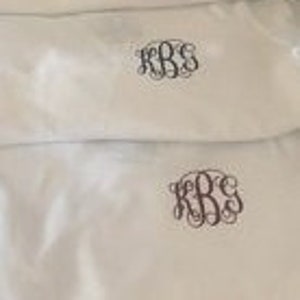 May include: Four white fabric tote bags with different colored embroidered monograms. The monograms are all the same design, but in different colors: pink, black, brown, and pink.