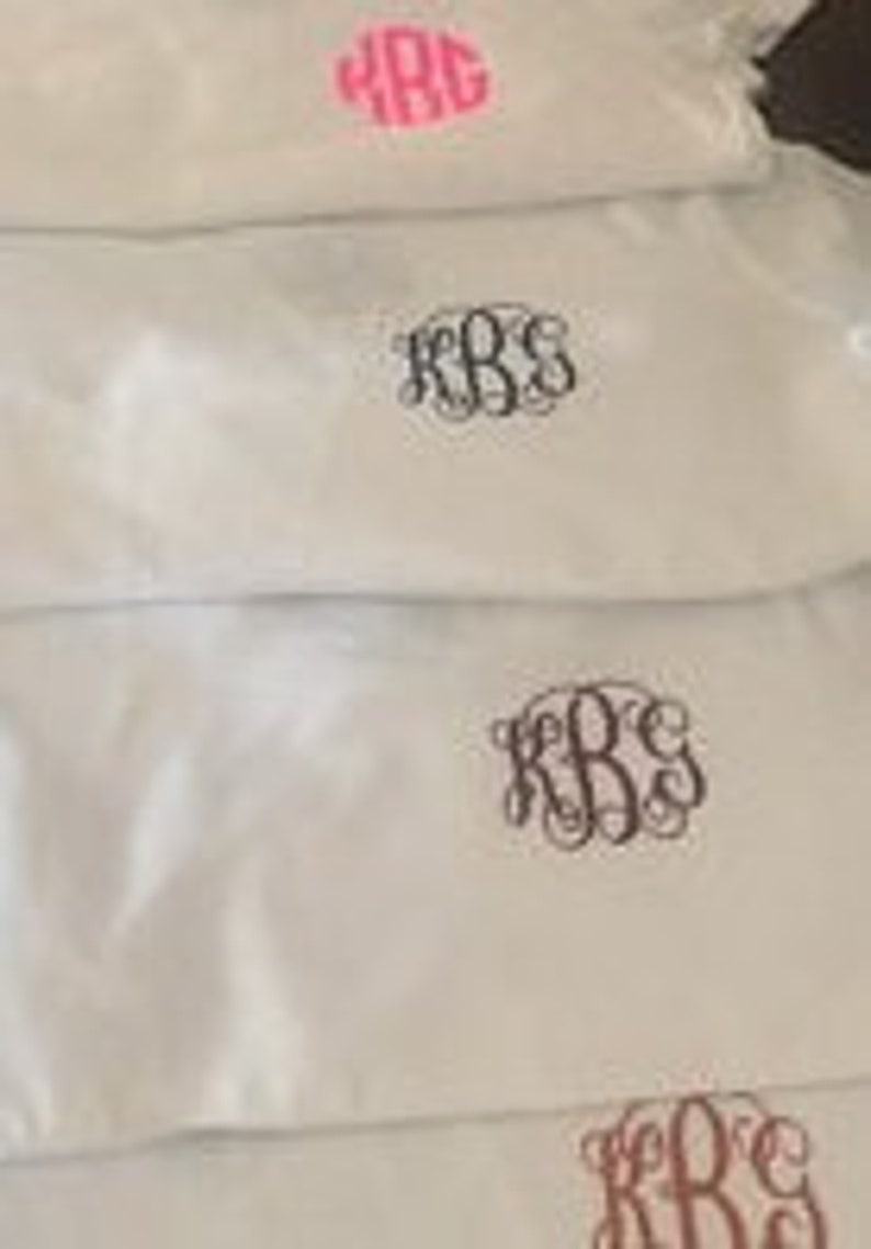 May include: Four white fabric tote bags with different colored embroidered monograms. The monograms are all the same design, but in different colors: pink, black, brown, and pink.