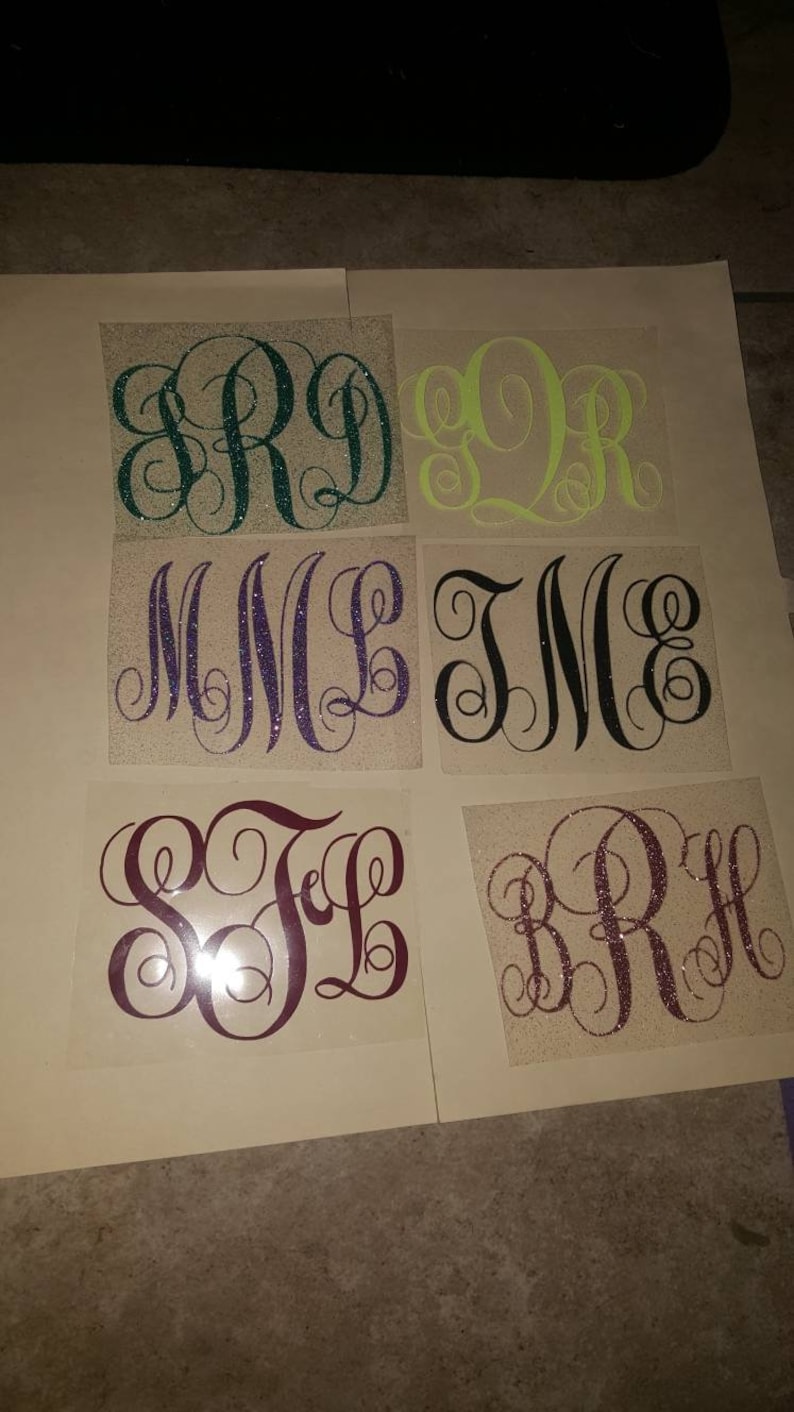 May include: Six different monogram designs on a white background. The monograms are made of glitter and are in different colors, including green, yellow, purple, black, and maroon.